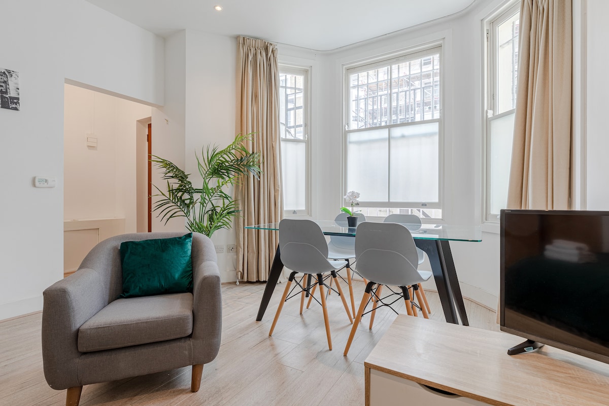 A light-filled living area features a glass dining table surrounded by four sleek chairs. A potted plant adds a touch of greenery, while a cozy armchair provides a comfortable seating option. Large windows with sheer curtains allow natural light to brighten the space.