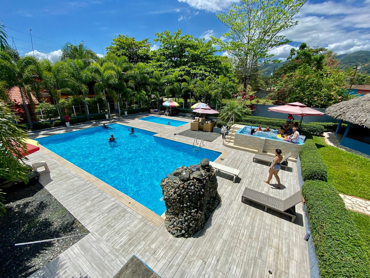 A spacious outdoor pool area is surrounded by tropical greenery, featuring a large pool for adults and a smaller pool for children. Sun loungers and umbrellas are positioned around the water. A jacuzzi is visible near the pool, offering a relaxed space for guests.
