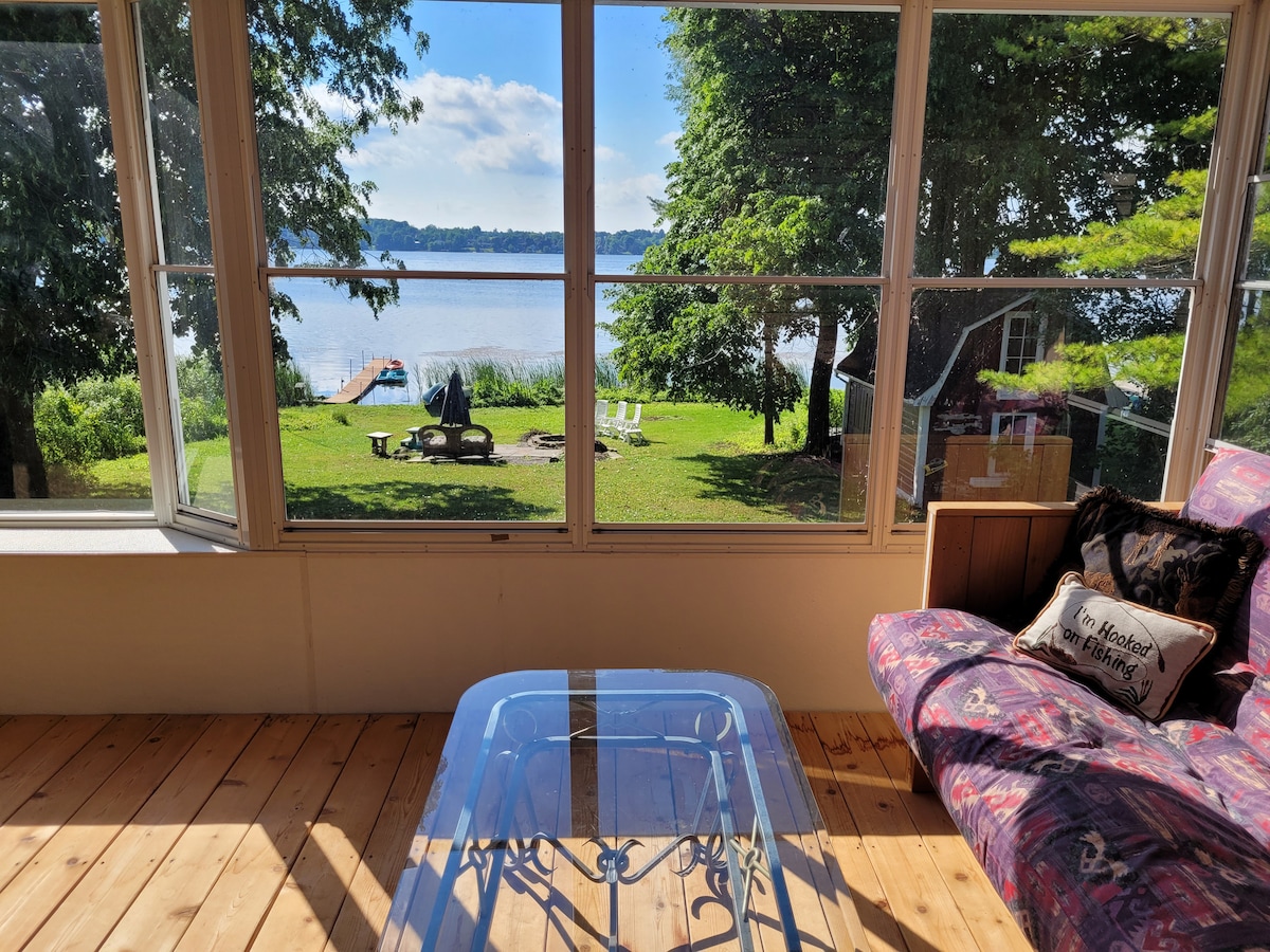 A sunroom flooded with natural light features a glass table and a cozy sofa, providing views of the serene waterfront and lush green lawn. Outside, a dock is visible, along with seating areas for relaxation by the water.