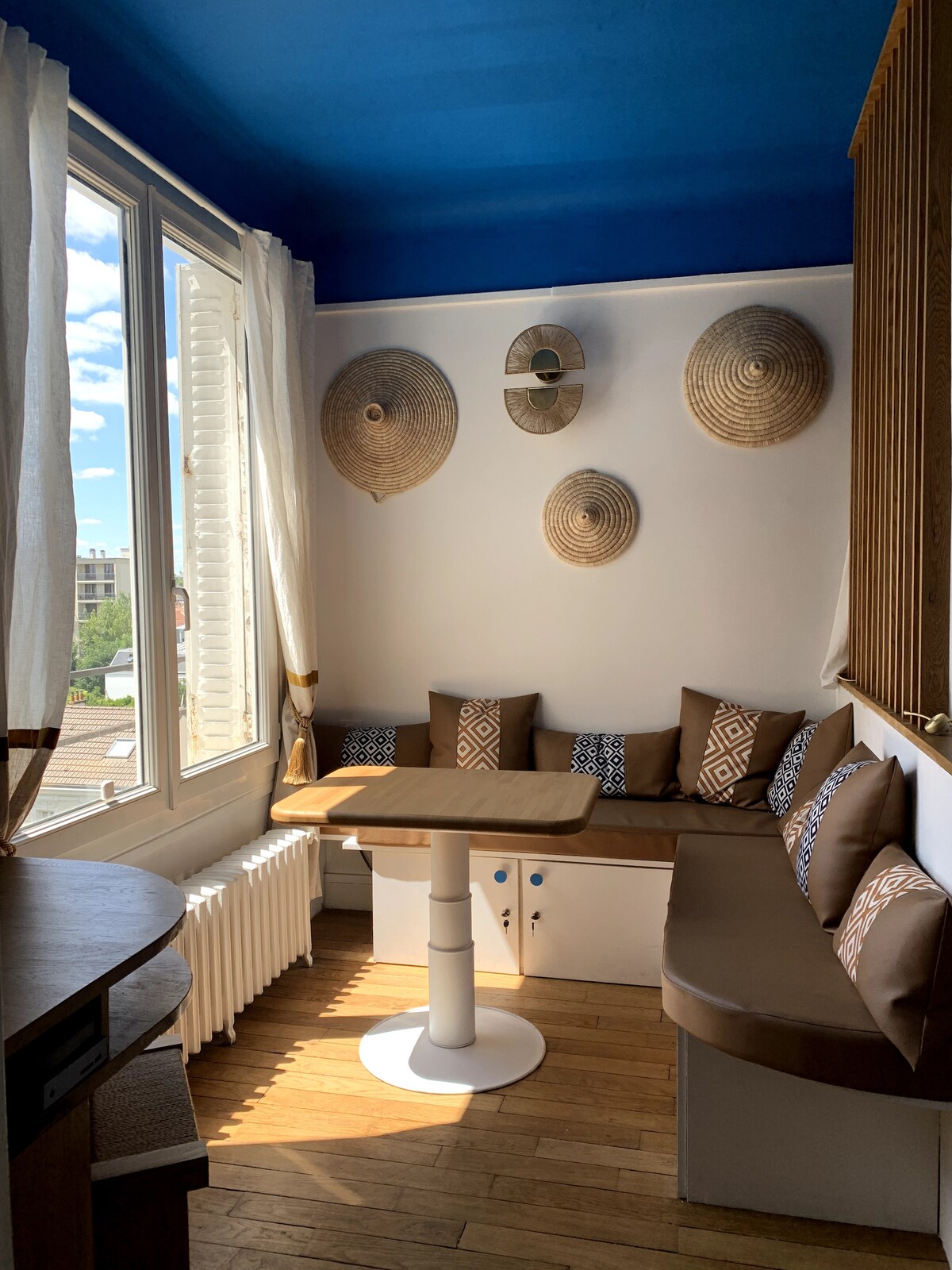 A bright corner of the living space is highlighted, featuring a round table with a white base. A brown cushioned bench with decorative pillows surrounds the table, and woven wall hangings add texture to the room. Large windows allow natural light to fill the area.