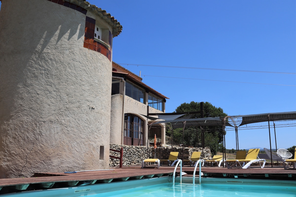A spacious swimming pool is surrounded by a wooden deck, featuring sun loungers positioned for relaxation. The unique architecture of a stone building is visible in the background, with clear blue skies overhead. The tranquil environment emphasizes the connection to nature.