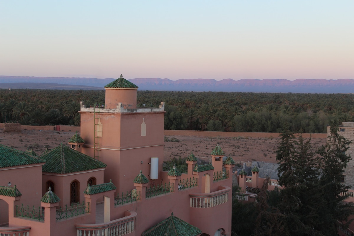 The image captures a view of a pink-hued building with distinctive green tiled roofs, complemented by surrounding lush palm trees and the vast desert landscape. In the background, mountains are softly outlined against a pastel sky, creating a calm and serene atmosphere.