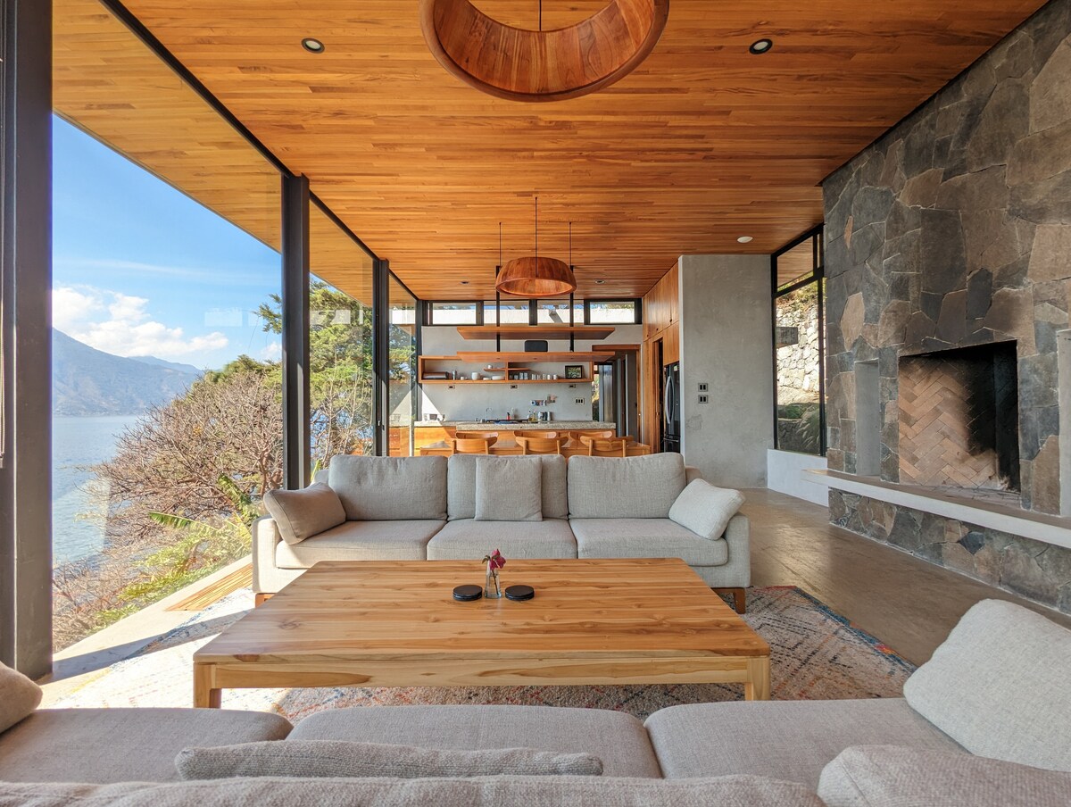The open-plan living area features large windows framing stunning lake views. A natural wooden coffee table is centered between light-colored sofas, inviting relaxation. The space includes a stone fireplace and sleek ceiling fixtures, highlighting the minimalist design in harmony with nature.
