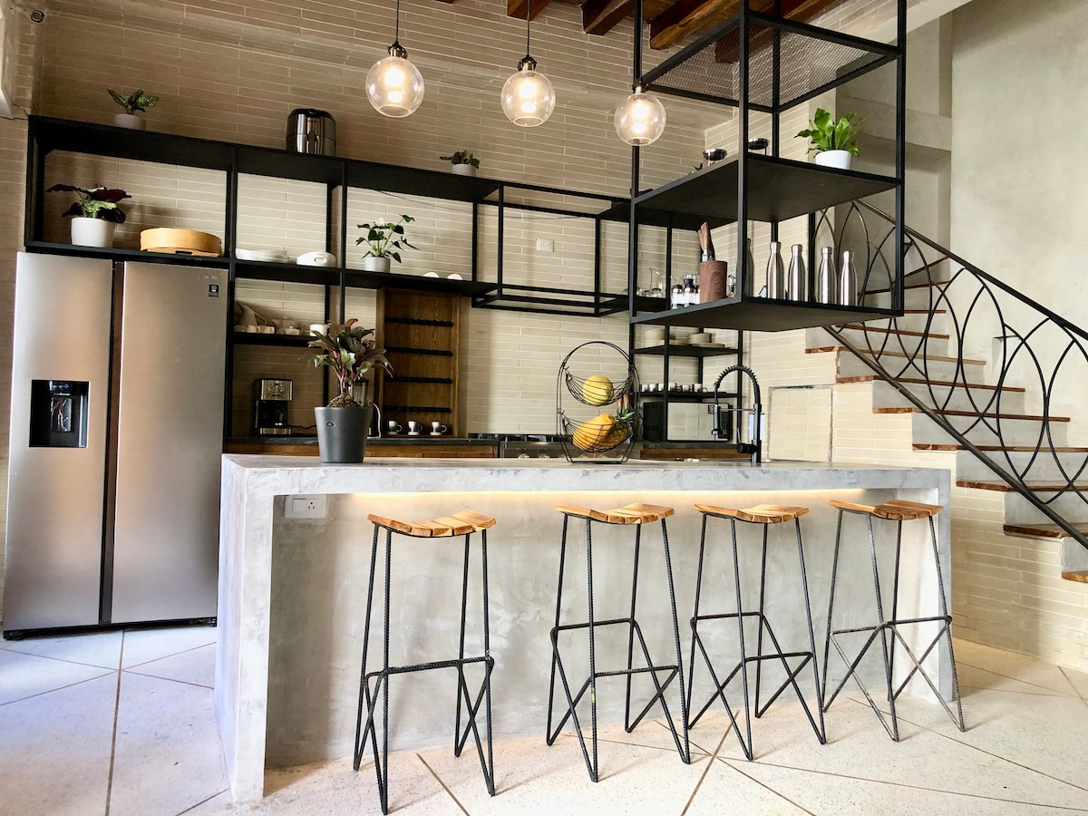 A modern kitchen is showcased with a sleek island featuring four high stools. A large refrigerator and black shelving units are visible, displaying decorative plants and kitchen items. Light fixtures hang above, and a staircase with an elegant railing leads to the upper level.