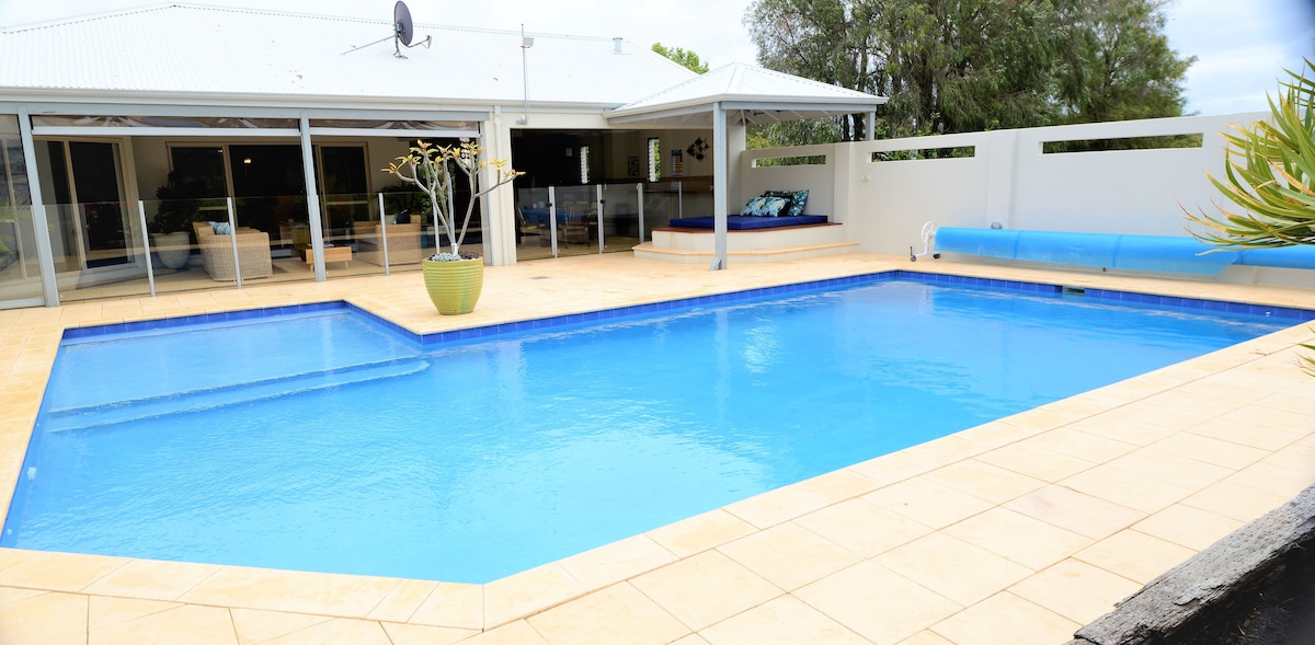 A spacious outdoor area surrounds a crystal-clear swimming pool, featuring a shallow section suitable for children. A covered seating area is situated nearby, providing shade and comfort. Lush greenery frames the scene, enhancing the inviting atmosphere of the backyard.