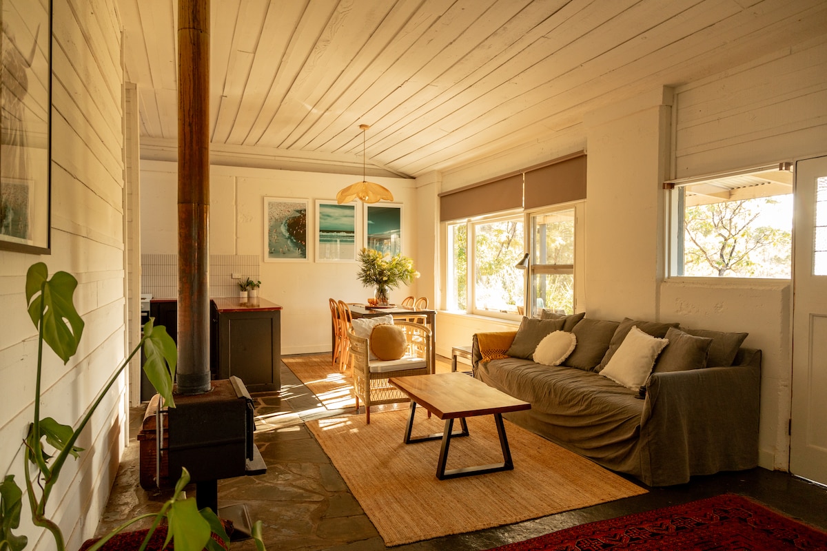 A cozy living area features a spacious sofa with decorative pillows, a wooden coffee table, and a dining table with wicker chairs. Sunlight enters through large windows, illuminating the space. Natural materials and neutral tones create a calm and welcoming environment.
