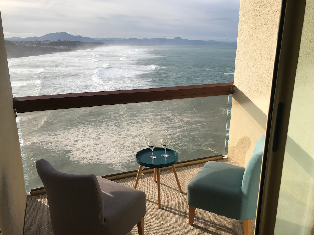 A covered balcony features two chairs positioned beside a small round table, displaying two glasses. The view showcases rolling ocean waves, with the distant mountains providing a serene backdrop.