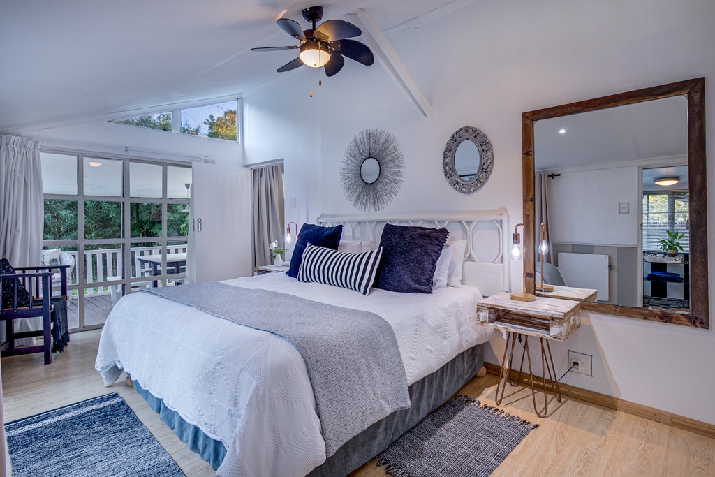 A spacious bedroom is equipped with a large bed adorned with decorative pillows and a soft blanket. Natural light enters through a large window, highlighting a rustic mirror and two round wall decor pieces. A cozy seating area is visible through the open doorway.