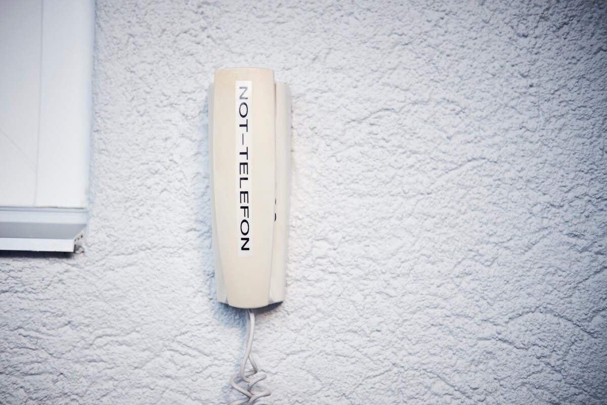 A beige emergency telephone is mounted on a textured white wall. The device is clearly labeled with the word 'NOT-Telefon,' providing a visual indication of its purpose. An electrical cord extends from the base, making connectivity evident.