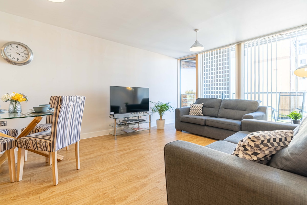 A modern living area features a large Smart TV on a glass stand, complemented by a grey sofa and an armchair. A dining table with striped chairs is set for four, while greenery adds a refreshing touch near the balcony doors with vertical blinds.