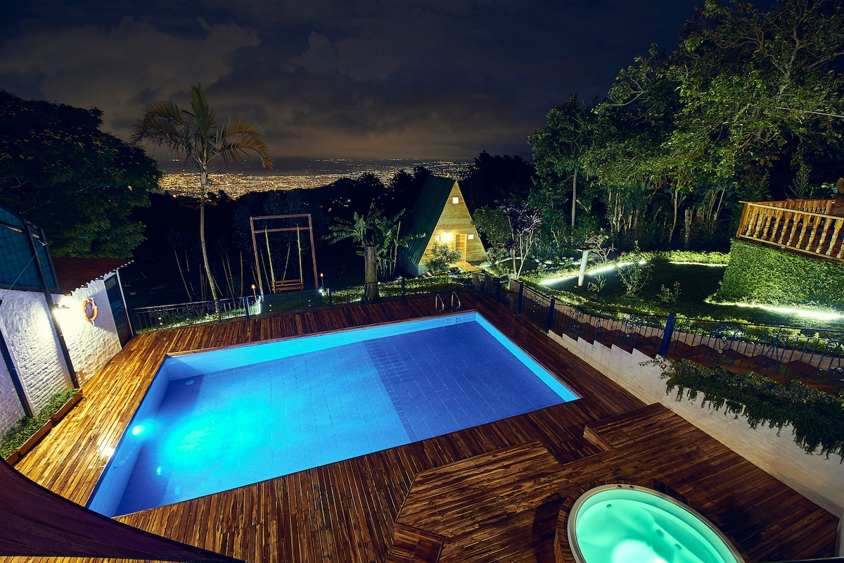 A tranquil scene showcases a lit swimming pool beside a heated Jacuzzi, both surrounded by wooden decking. Lush greenery frames the space, while twinkling city lights are visible in the background, creating a serene evening atmosphere.