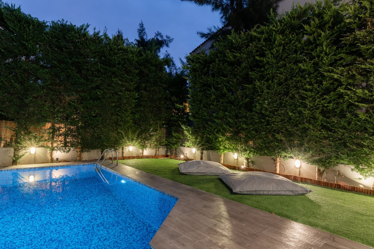 A private pool area is surrounded by lush greenery, creating a serene outdoor space. The shining blue water of the pool is complemented by comfortable oversized cushions on the deck, with subtle ground lighting illuminating the perimeter.