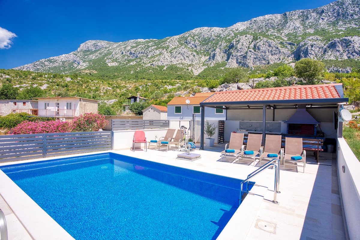 A large, inviting pool is framed by a spacious deck and complemented by comfortable loungers. The backdrop features a mountain landscape, providing a serene view. A covered area is equipped with a grill, ideal for outdoor cooking and gatherings.