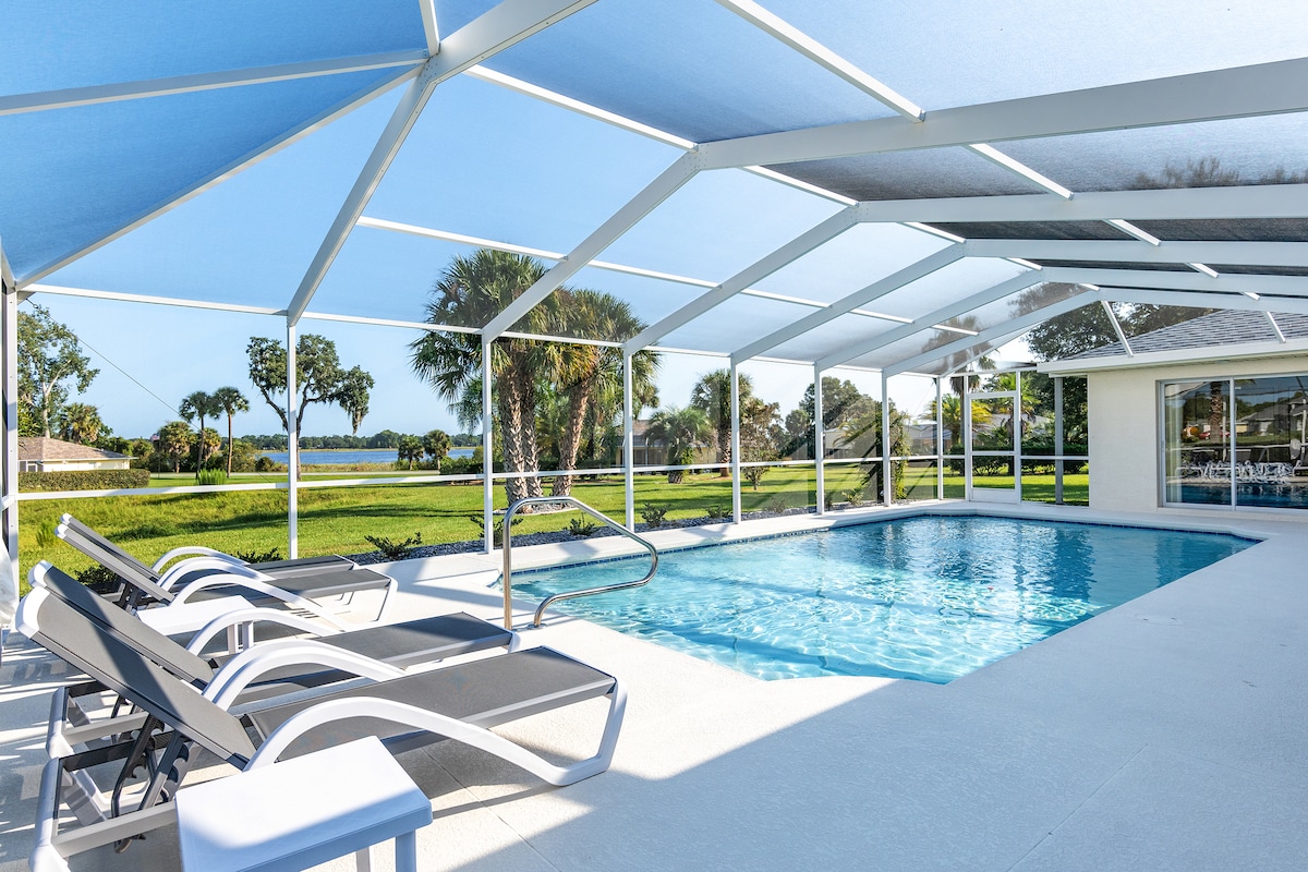 The outdoor pool area features a large screened enclosure, allowing natural light while providing shade. Lounge chairs are positioned around the pool's edge. Lush greenery is visible in the surrounding landscape, offering a serene backdrop for relaxation.