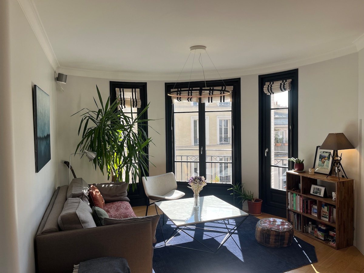 A bright living area features a comfortable sofa and a cozy armchair, arranged around a marble-topped coffee table. Large windows with black frames allow natural light to fill the space, complemented by a flourishing indoor plant. A soft rug anchors the seating area.