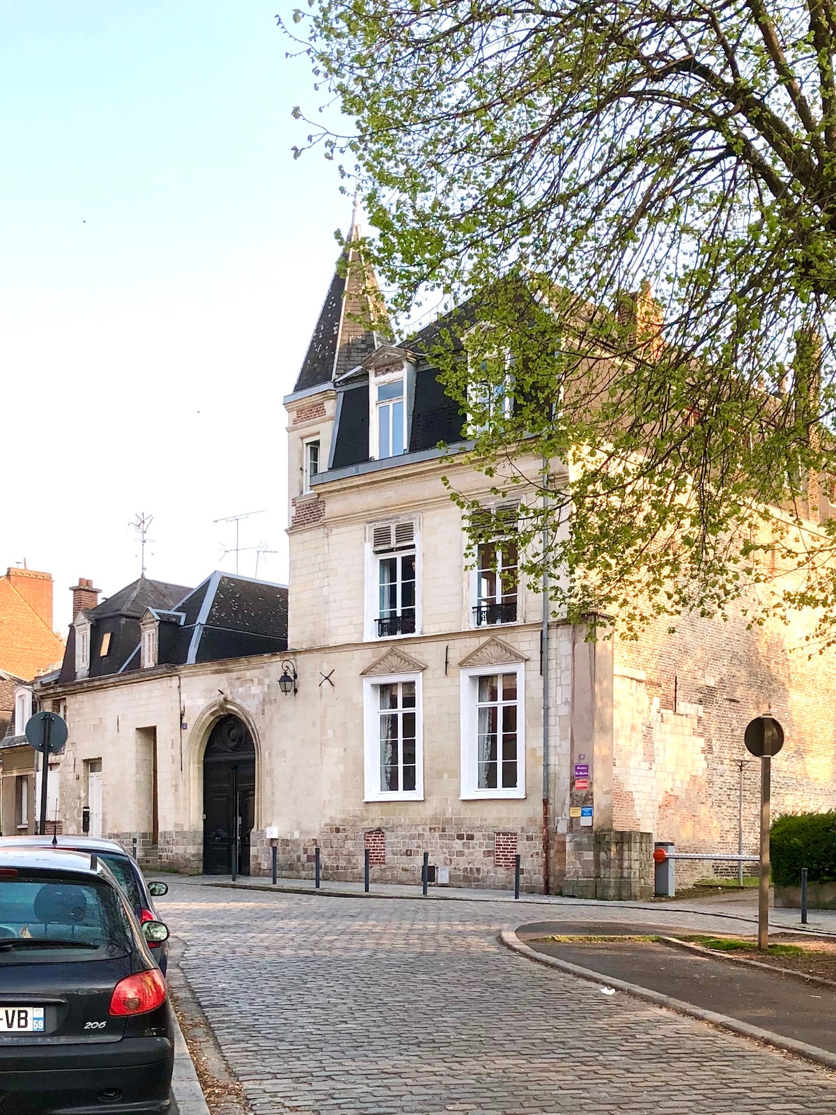 A charming building characterized by a blend of stone and traditional architecture is depicted from the street view. Large windows and a steeply pitched roof highlight its unique design, surrounded by greenery and cobblestone streets that enhance the inviting atmosphere of the neighborhood.