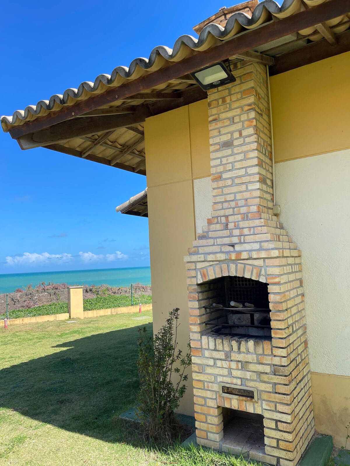 An outdoor grill is situated beside the exterior wall of the chalet, crafted from light-colored brick. The background captures a view of the ocean under a clear blue sky, with greenery surrounding the area.