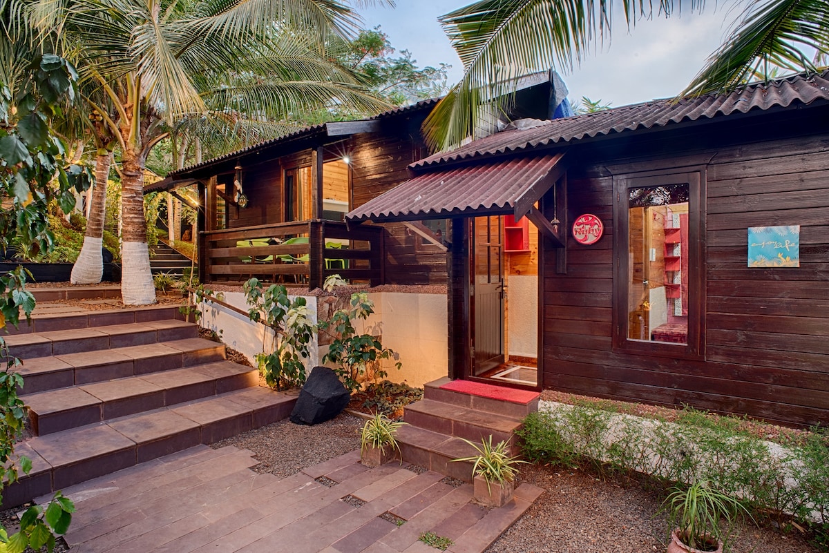 A charming exterior of the villa is showcased, featuring a warm wooden facade complemented by lush tropical plants. Steps lead up to the entrance, with a welcoming patio area adorned with potted greenery, creating an inviting atmosphere amidst the natural surroundings.