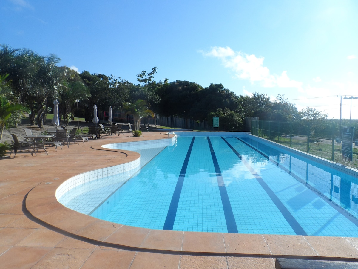 A spacious outdoor pool is visible, featuring clear blue water and neatly arranged lanes. Surrounding the pool are lounge chairs that provide ample seating. Lush greenery lines the background, contributing to a serene atmosphere.