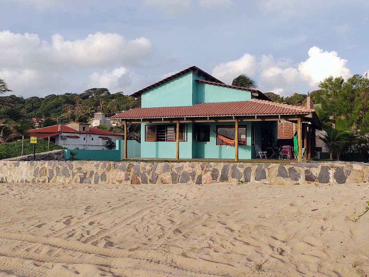 A beach house is depicted, featuring a colorful exterior in shades of blue and red. The structure sits near a sandy area, with a stone wall in the foreground and a porch adorned with chairs and a hammock. Lush vegetation and hills line the background.