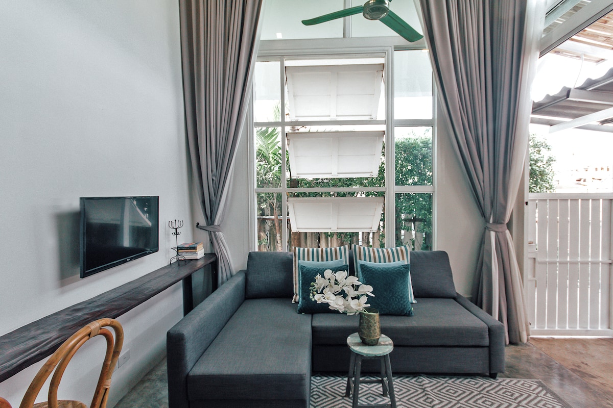 A spacious living area features a gray sectional sofa adorned with turquoise throw pillows. A small coffee table holds decorative flowers, while a flat-screen TV is mounted on the wall. Large windows allow natural light to fill the space, complemented by light drapes and a ceiling fan.