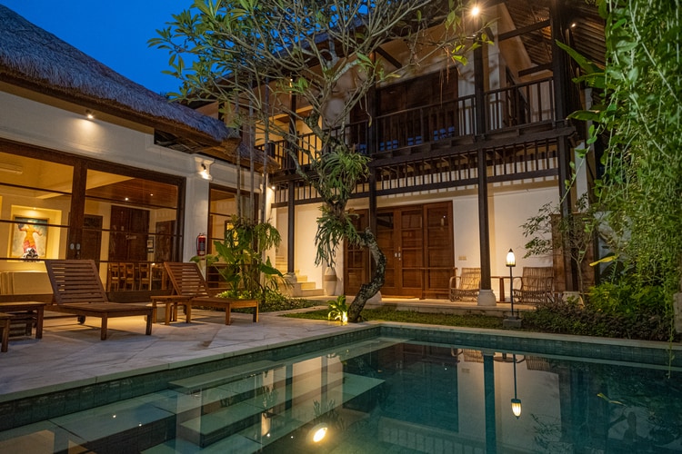 The villa is showcased in a tranquil evening setting, featuring a private swimming pool reflecting the surrounding greenery. Sun loungers are positioned beside the pool, while the two-story structure, with wooden balconies, is illuminated softly, blending seamlessly with the lush landscape.