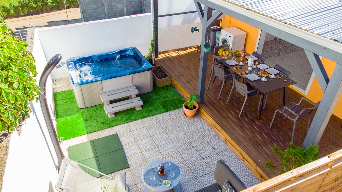 An outdoor space features a hot tub and an elegant dining area beneath a covered terrace. A table is set for a meal, surrounded by sleek chairs. Lush planters and green grass add vibrant touches to the relaxation area, which is complemented by a wooden deck.