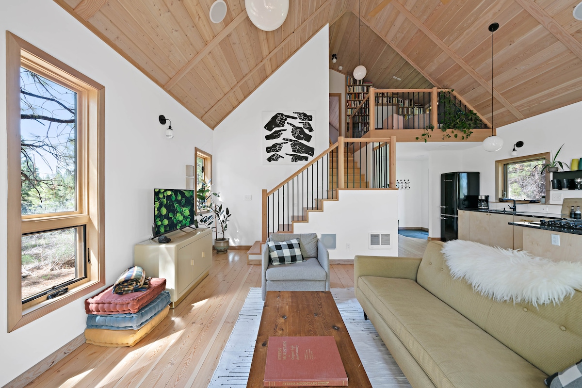 An open living area features a modern sofa with a faux fur throw, accompanied by a wooden coffee table. Natural light enters through large windows, highlighting the wooden ceiling and staircase that leads to a loft. A television and plants are visible in the room.