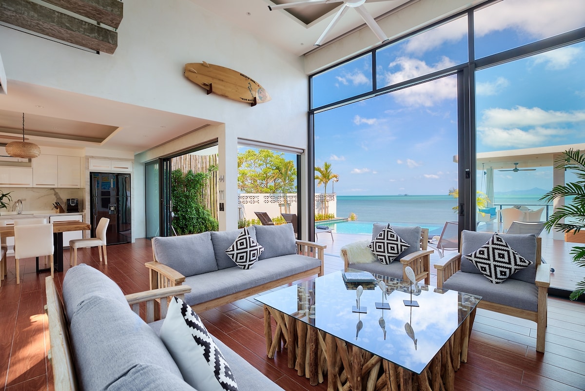 The open-plan living area features large glass doors that offer views of the sea. A wooden coffee table is surrounded by comfortable seating, with decorative cushions adding contrast. Light pours in from the expansive windows, highlighting the natural materials used throughout the space.