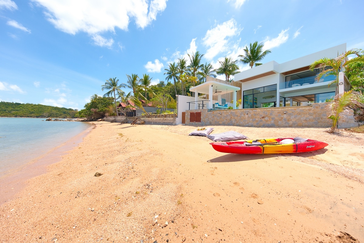 A sandy beach extends in front of the villa, with gentle waves lapping at the shore. Palm trees provide shade, and a kayak rests on the sand, suggesting opportunities for water activities. The modern villa features large glass windows, blending seamlessly with the coastal surroundings.