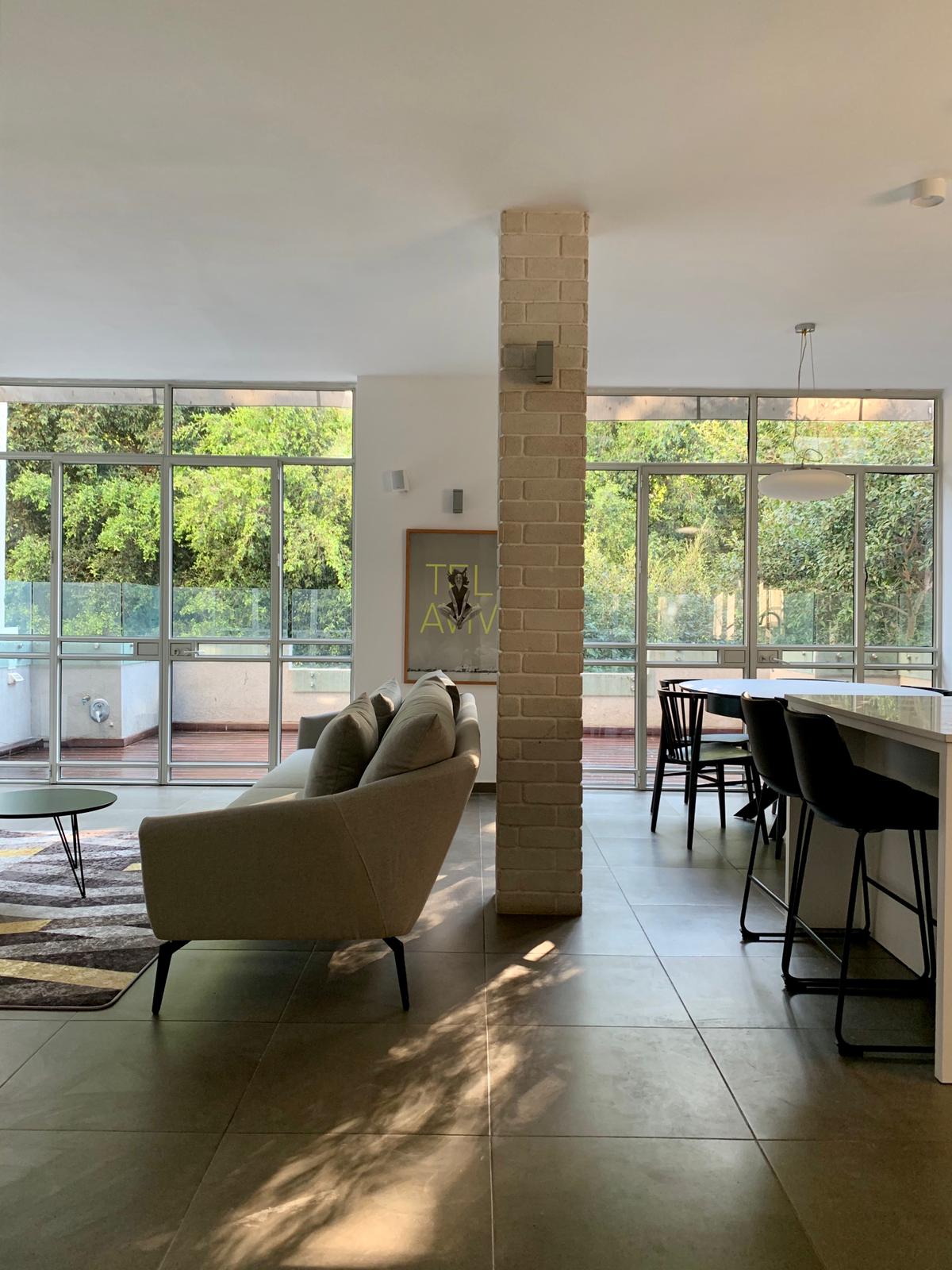 A bright and airy living space features large windows allowing natural light to fill the room. A modern sofa sits in the foreground, accompanied by a patterned area rug. A kitchen counter with stools is visible, offering a seamless flow between dining and relaxation areas.