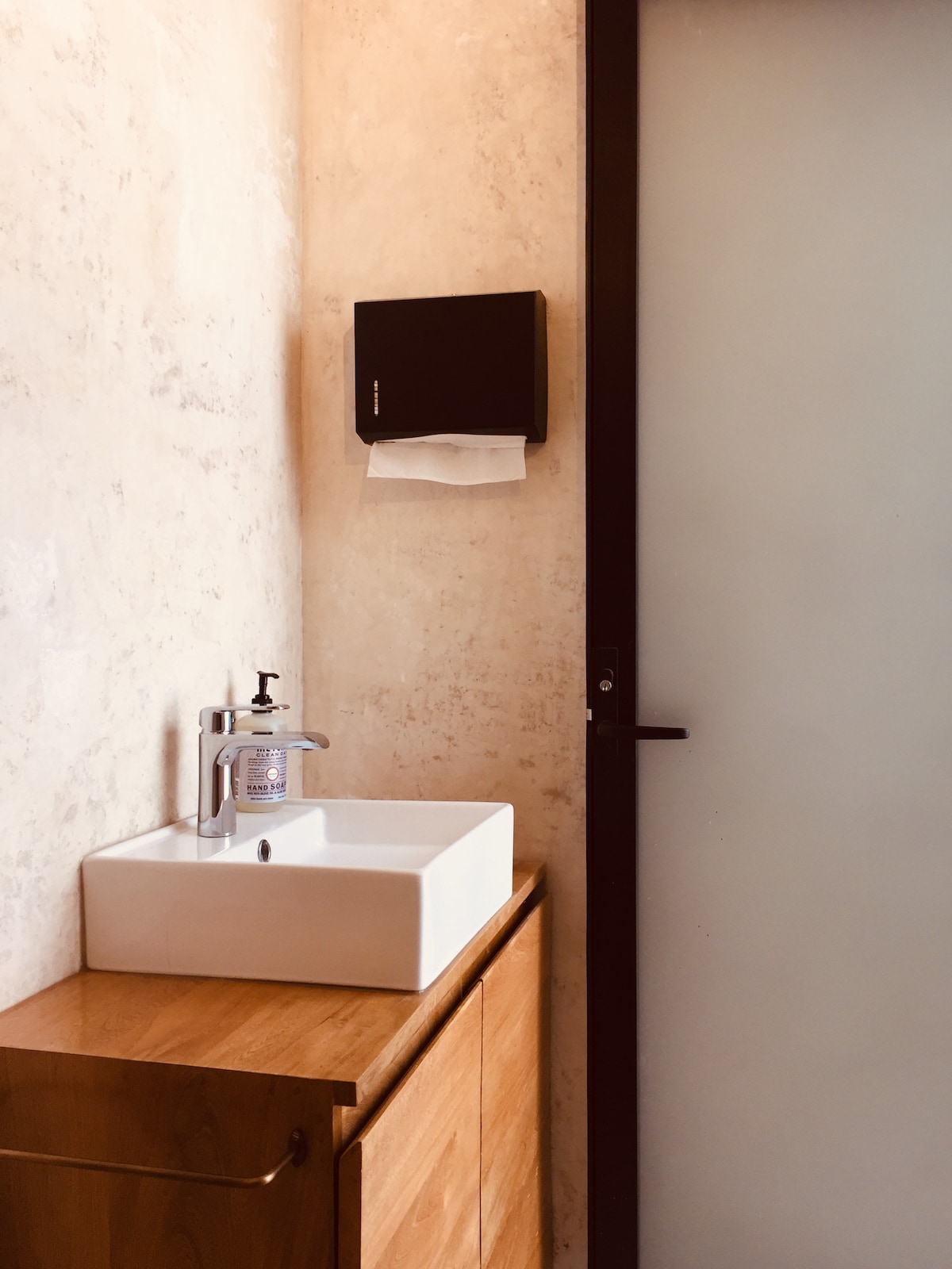 A clean and modern bathroom space is showcased, featuring a minimalist white sink atop a wooden cabinet. A soap dispenser is positioned next to the sink, while a frosted glass door adds privacy to the area.