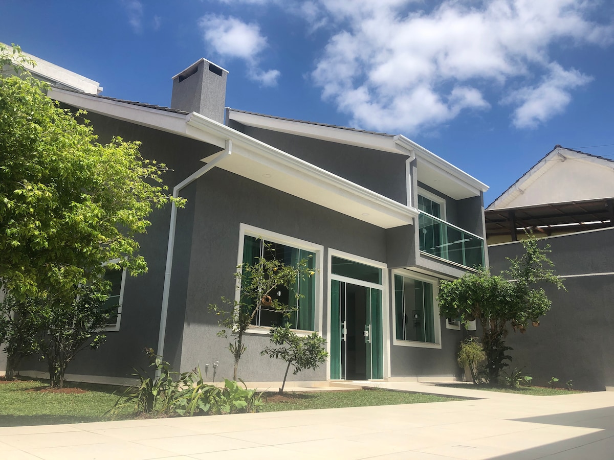 The exterior of the house features a modern design with large glass windows allowing natural light. Lush green plants and two trees frame the area, complementing the gray façade under a clear blue sky.