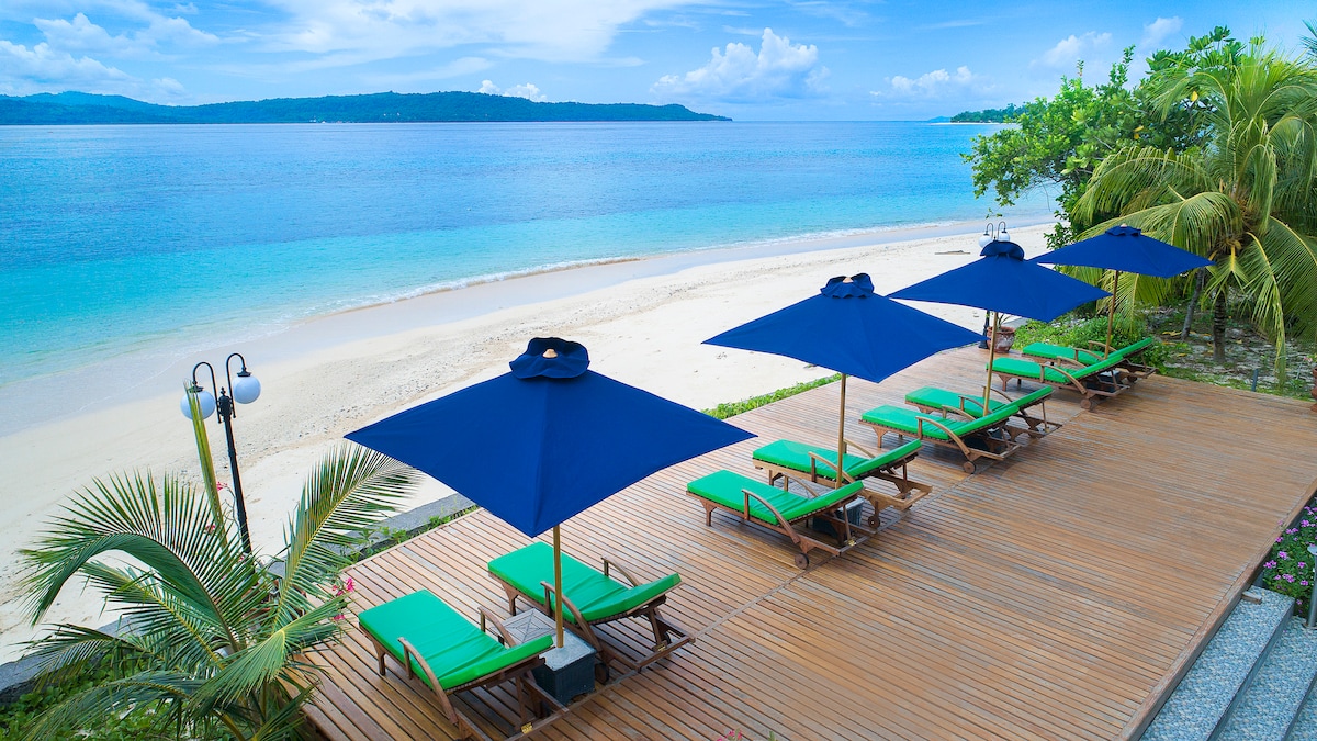 Lounge chairs are arranged under large blue umbrellas on a wooden deck, overlooking a serene private beach. The beach extends along calm, clear waters, framed by lush greenery and distant islands against a bright sky.