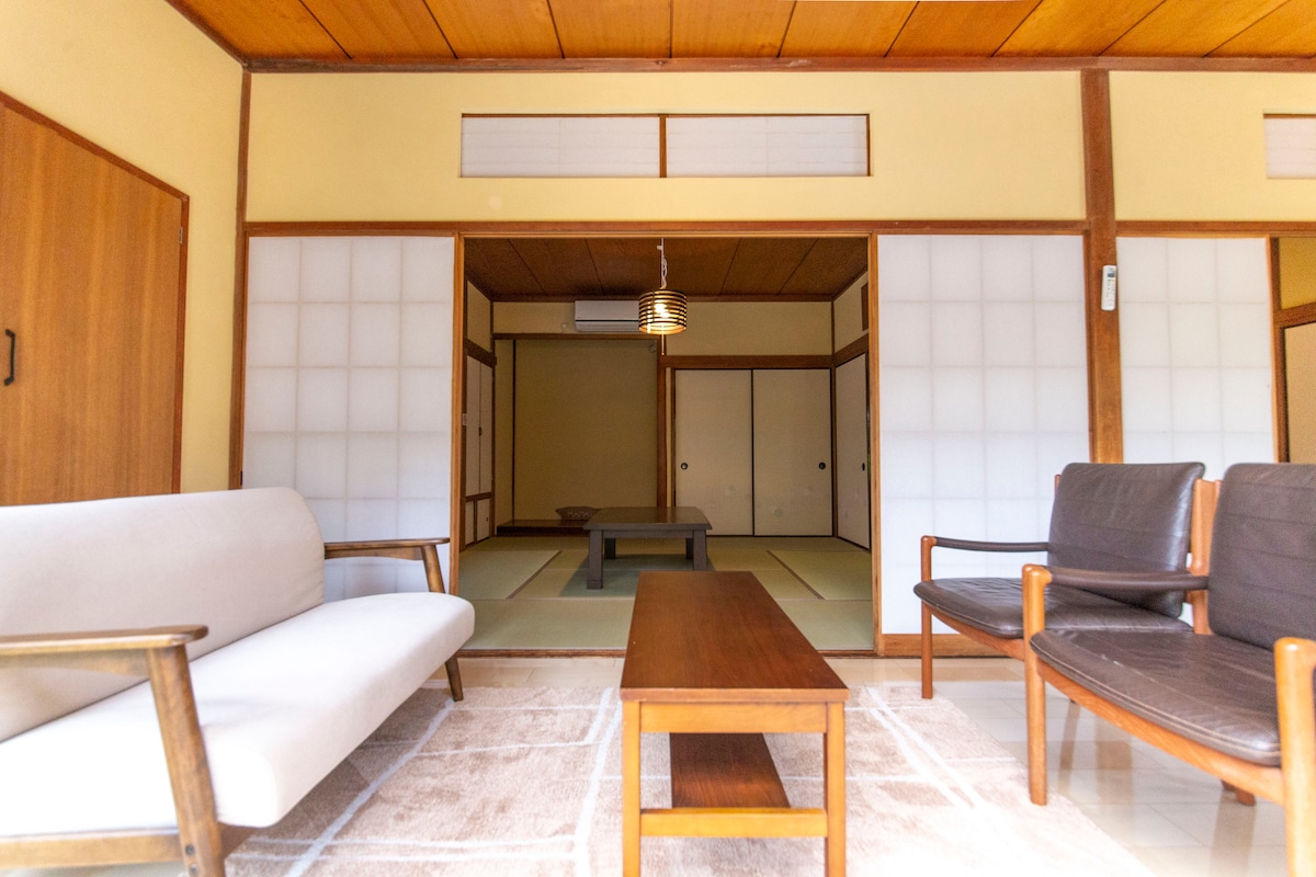 A light-filled living area features a simple white sofa and two dark wooden chairs surrounding a low wooden table. The backdrop consists of sliding shoji doors, with traditional tatami flooring visible in the adjacent room, showcasing the essence of a Japanese home.