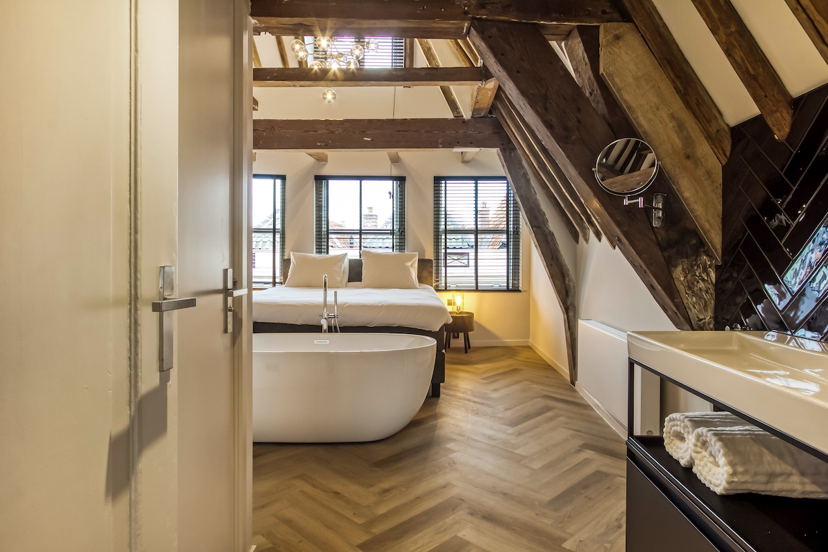An open layout showcases a spacious bed beneath sloped wooden beams. Natural light filters through multiple windows, highlighting the sleek freestanding bathtub nearby. A modern sink area accompanies a neatly arranged towel, creating a harmonious blend of contemporary design with historic elements.