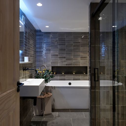 A modern bathroom is featured, showcasing sleek gray tiles on the walls and floor. A freestanding tub is positioned against a darker tile backdrop, accompanied by a minimalist sink on a wooden base. Soft lighting highlights the clean lines and tranquil atmosphere.