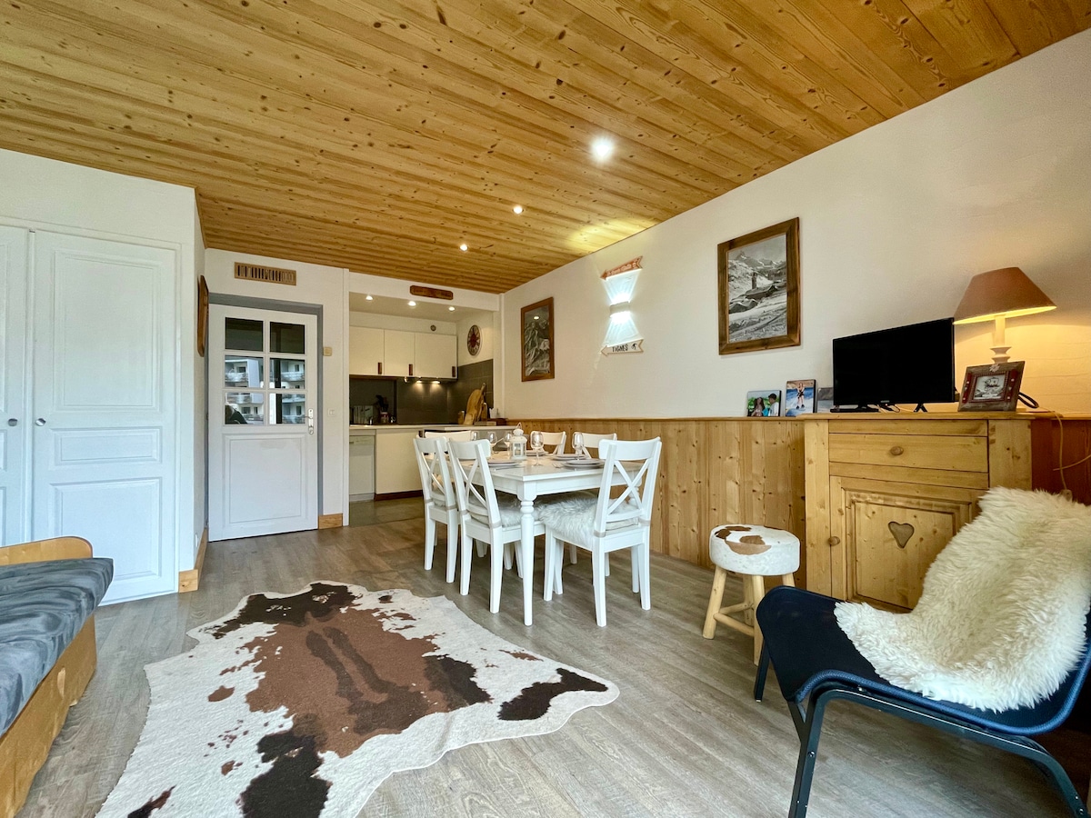 The inviting living area features a wooden ceiling and a mix of textures with a soft rug and cozy seating options. A dining table with white chairs is set for meals, while an open kitchen can be seen in the background, providing a seamless flow in the space.