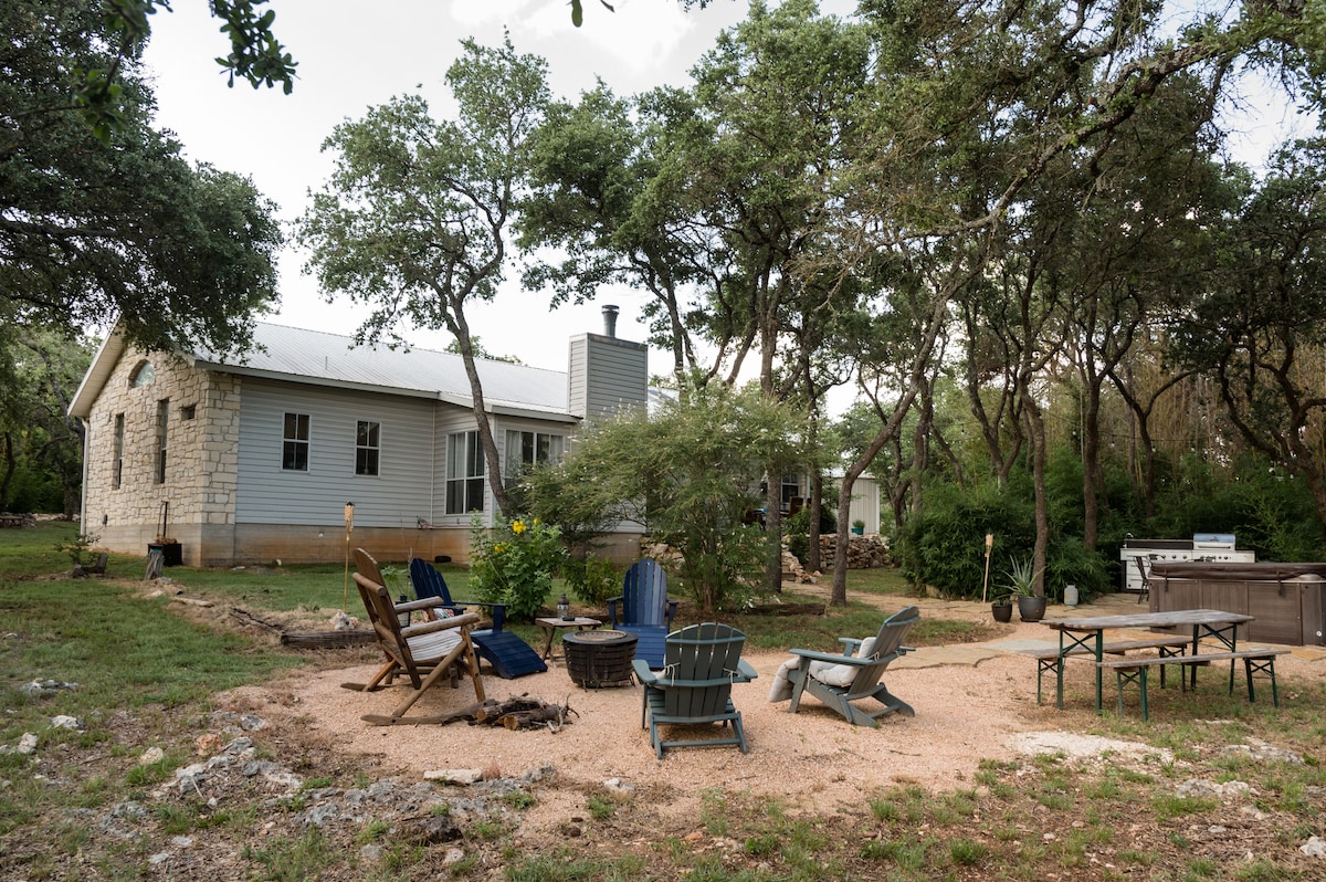 An outdoor gathering area is surrounded by lush oak trees, featuring a fire pit at the center. Several chairs, including wooden adirondack and a picnic table, are arranged on a gravel surface, creating an inviting atmosphere for relaxation. A hot tub is positioned nearby, blending with the natural setting.