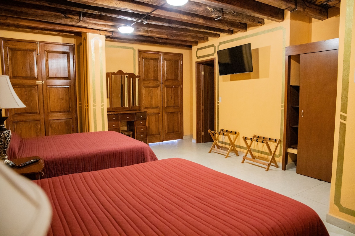 The room features two spacious beds with red bedspreads, complemented by wooden beams on the ceiling. A wooden dresser with a mirror is positioned against the wall, along with a wall-mounted television. Two folding stools are placed near the beds, providing additional seating.