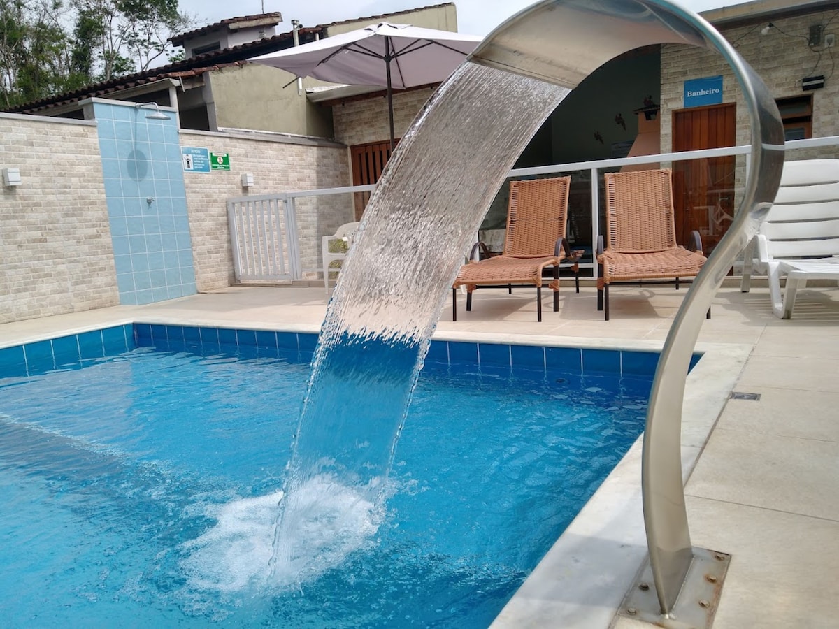 A swimming pool is featured with a waterfall spout, creating a gentle cascade into the clear water. Comfortable lounge chairs are positioned nearby, accompanied by an umbrella for shade. The area is surrounded by a sleek stone wall, enhancing the serene environment.