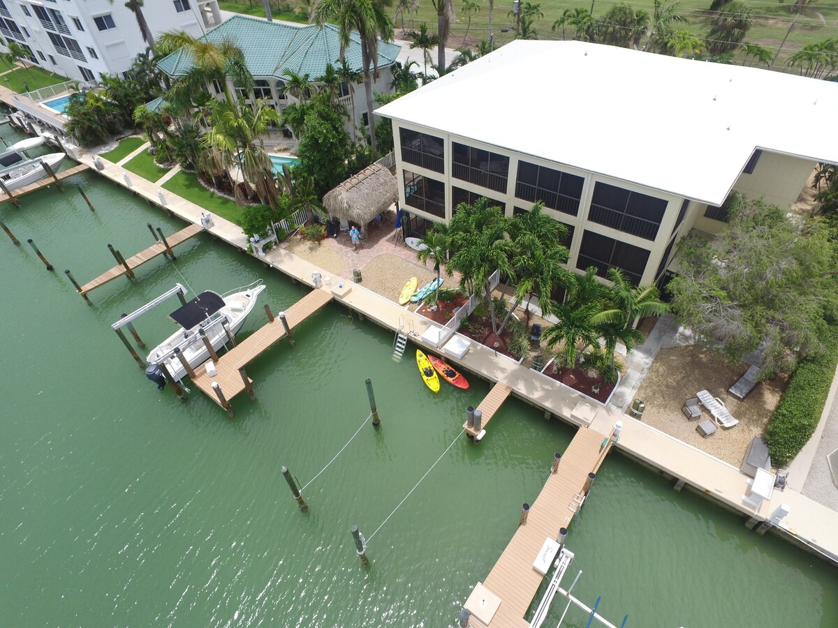 An aerial view captures the property adjacent to the water, with docks accommodating boats. A Tiki hut is nestled among palm trees, while kayaks are parked nearby on the shore. The house features expansive screened-in balconies, providing a view of the surrounding greenery and docks.