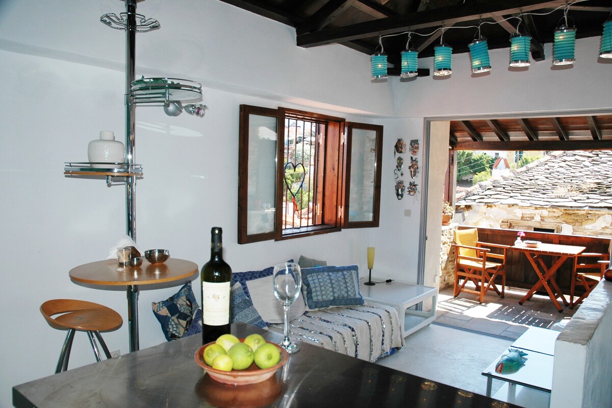 An open-plan living space features a seating area with a cozy divan and decorative pillows. A small dining table is present with stools, while a modern kitchen counter showcases a wine bottle and green apples. Natural light streams through the windows, illuminating the rustic decor.