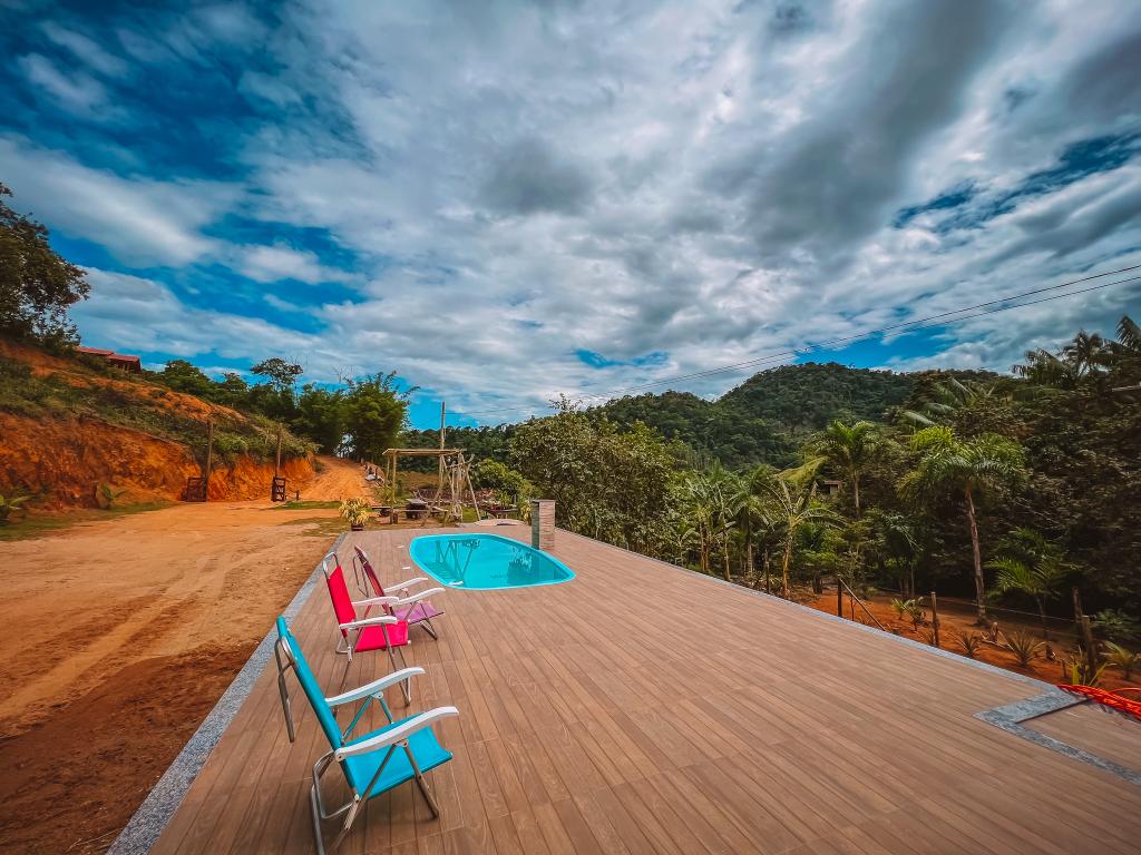 A wooden deck features a small, inviting pool surrounded by lounge chairs in pink and blue. Lush greenery and gentle hills are visible in the background under a cloudy sky, offering a serene outdoor retreat.