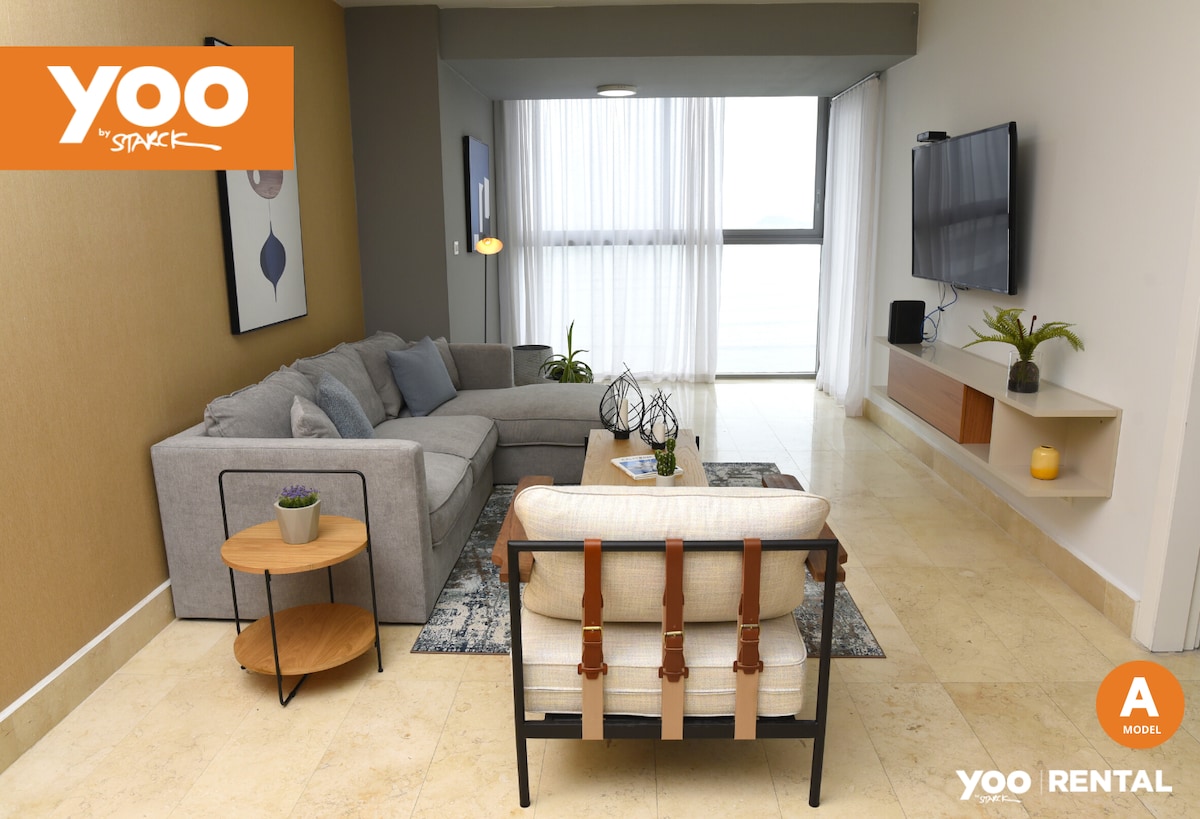 A spacious living area is presented, featuring a modern gray sectional sofa and a light-colored armchair. Natural light flows through large windows covered by sheer curtains. A wooden coffee table sits in the center, accompanied by a small side table and a television mounted on the wall.