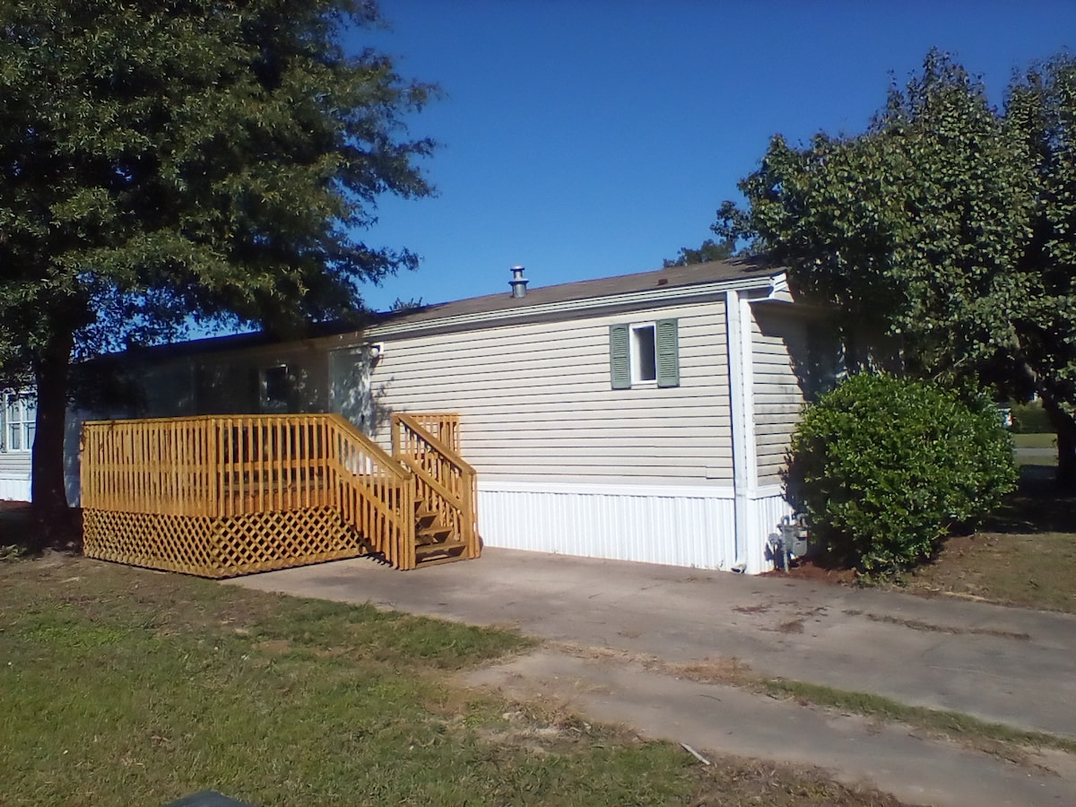 A remodeled home with a spacious wooden deck is presented, featuring a ramp for accessibility. The exterior showcases neutral siding and a well-maintained yard with greenery. Large windows allow for natural light, ensuring a welcoming appearance.