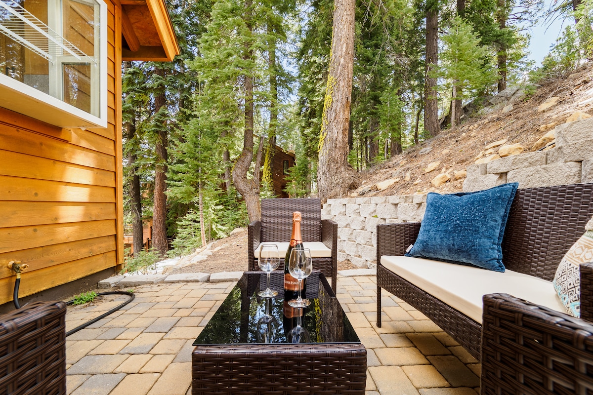 A cozy outdoor seating area features two wicker chairs and a comfortable sofa, arranged around a low glass-top table. Surrounding greenery includes tall trees and stone walls, creating a tranquil environment for relaxation.