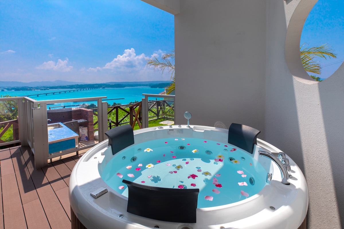 A spacious outdoor jacuzzi is positioned on the deck, with water adorned by colorful flower petals. Views of a sparkling sea and the distant bridge are visible in the background, framed by a bright blue sky. The serene setting is complemented by surrounding railings.