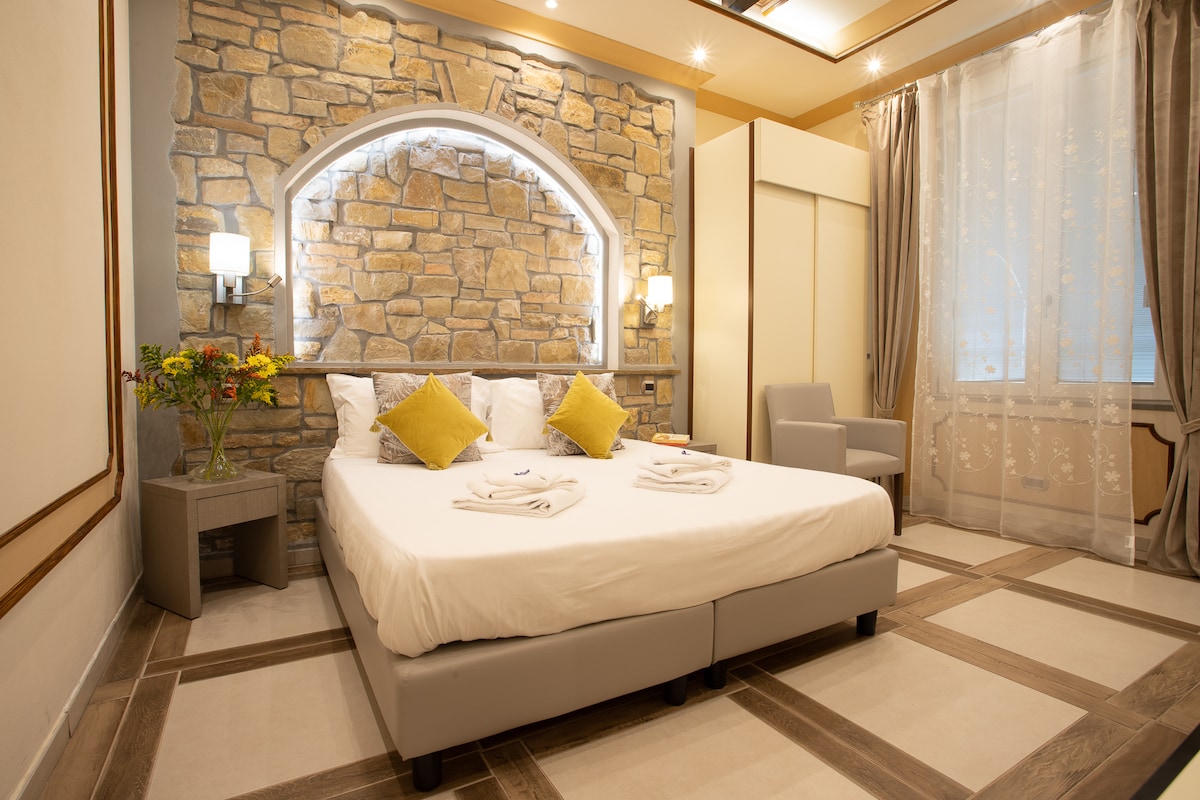 A comfort-oriented sleeping area features a spacious bed with white linens and two yellow accent pillows. A stone wall with an arched nook adds texture, while soft lighting creates a serene ambiance. A side table holds fresh flowers, and light drapes frame a window.