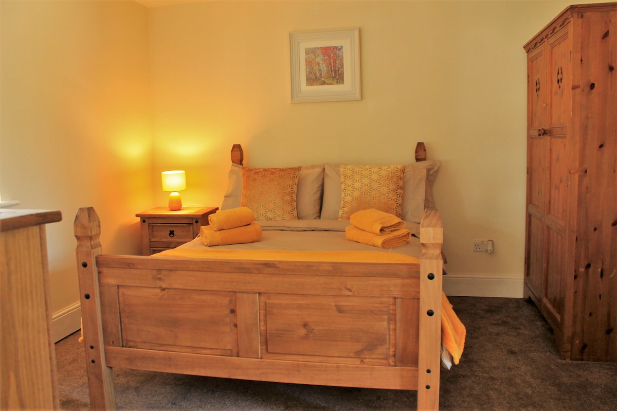 A warm and inviting bedroom is featured, showcasing a wooden bed with an additional spacious sleeping area. Freshly folded towels are neatly arranged on the bed. Soft lighting illuminates the room, complemented by wooden furniture and a tranquil wall art piece.
